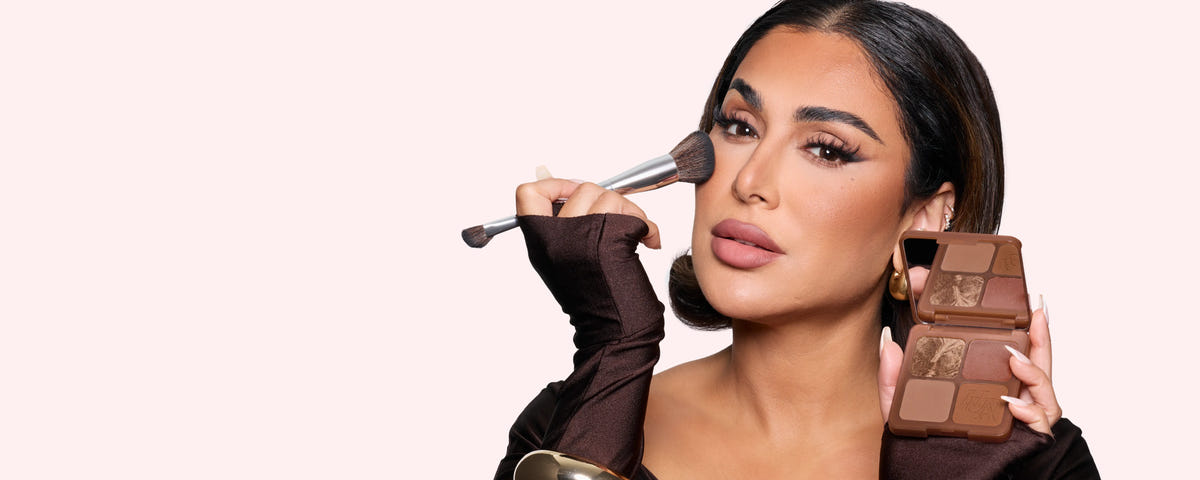 Huda Beauty Bronze Filter Palette promotional banner of founder Huda Kattan applying bronzer with a brush and holding the open palette against a pale pink background