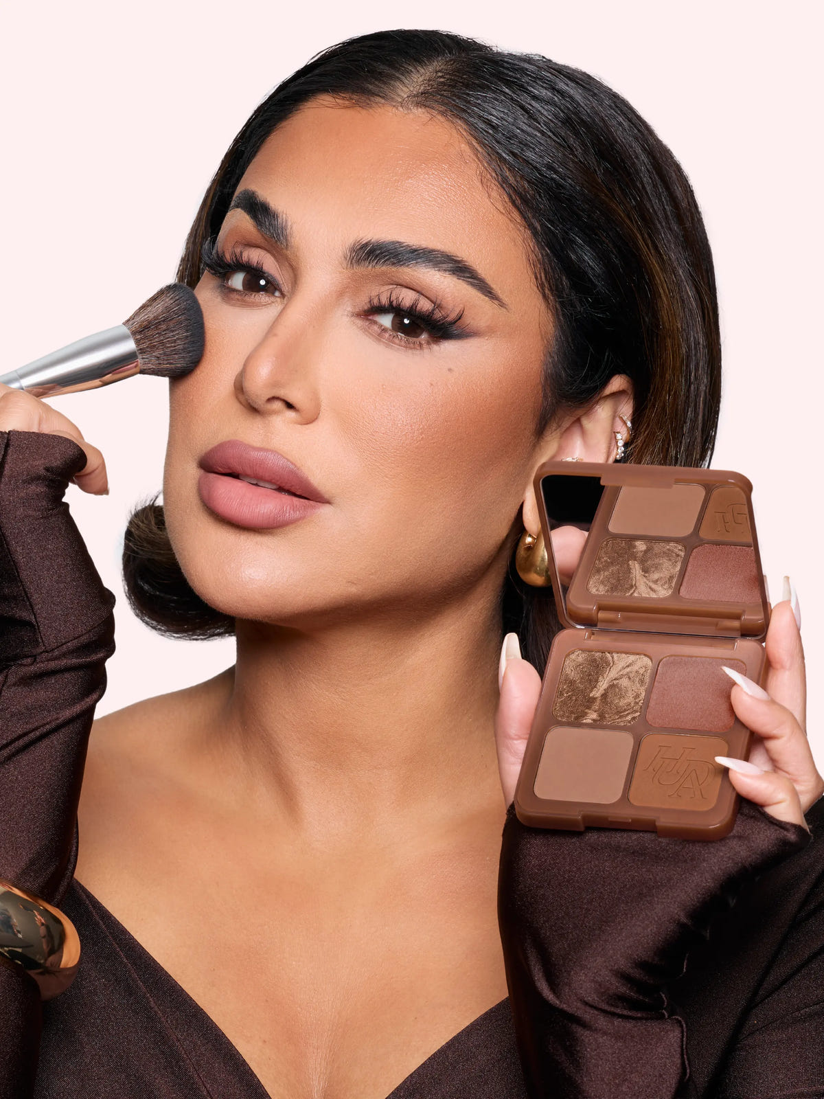 Huda Beauty Bronze Filter Palette promotional banner of founder Huda Kattan applying bronzer with a brush and holding the open palette against a pale pink background