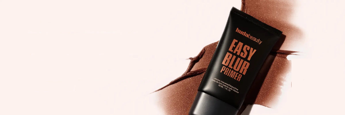 Huda Beauty Easy Blur Primer tube lying on a bronze product smear against a soft pink background