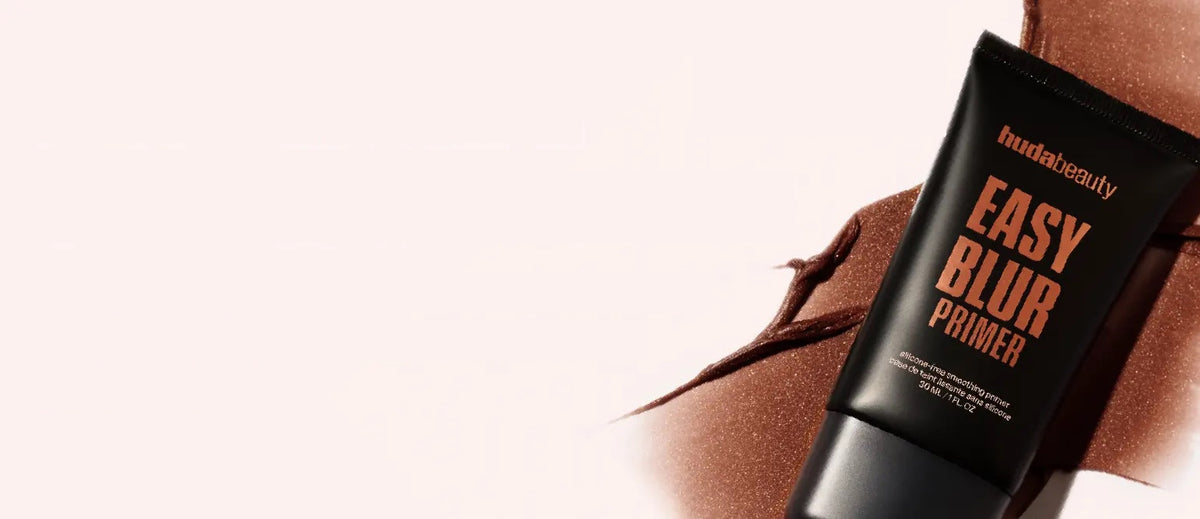 Huda Beauty Easy Blur Primer tube lying on a bronze product smear against a soft pink background