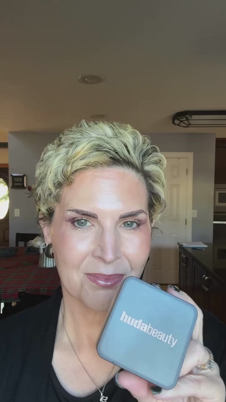 Woman bakes under her eyes with Easy Bake loose powder, presses it in with a small brush, then sweeps off the excess. 